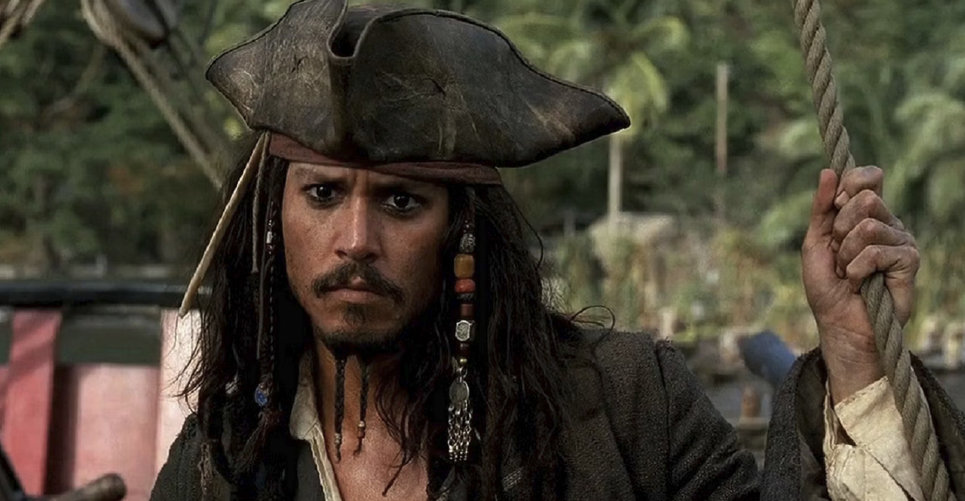 Johnny Depp as Jack Sparrow, a pirate with long hair, a tricorne hat, and beads in his beard stands holding a rope, with trees in the background in Pirates of the Caribbean.