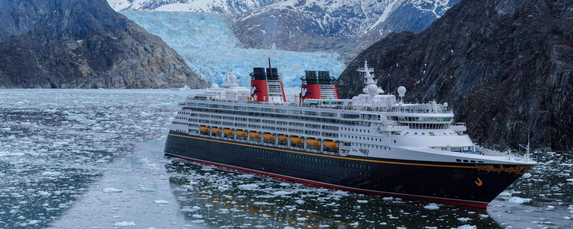 A Disney Wonder Alaska Cruise ship navigates icy waters near a glacier, surrounded by rocky mountains.