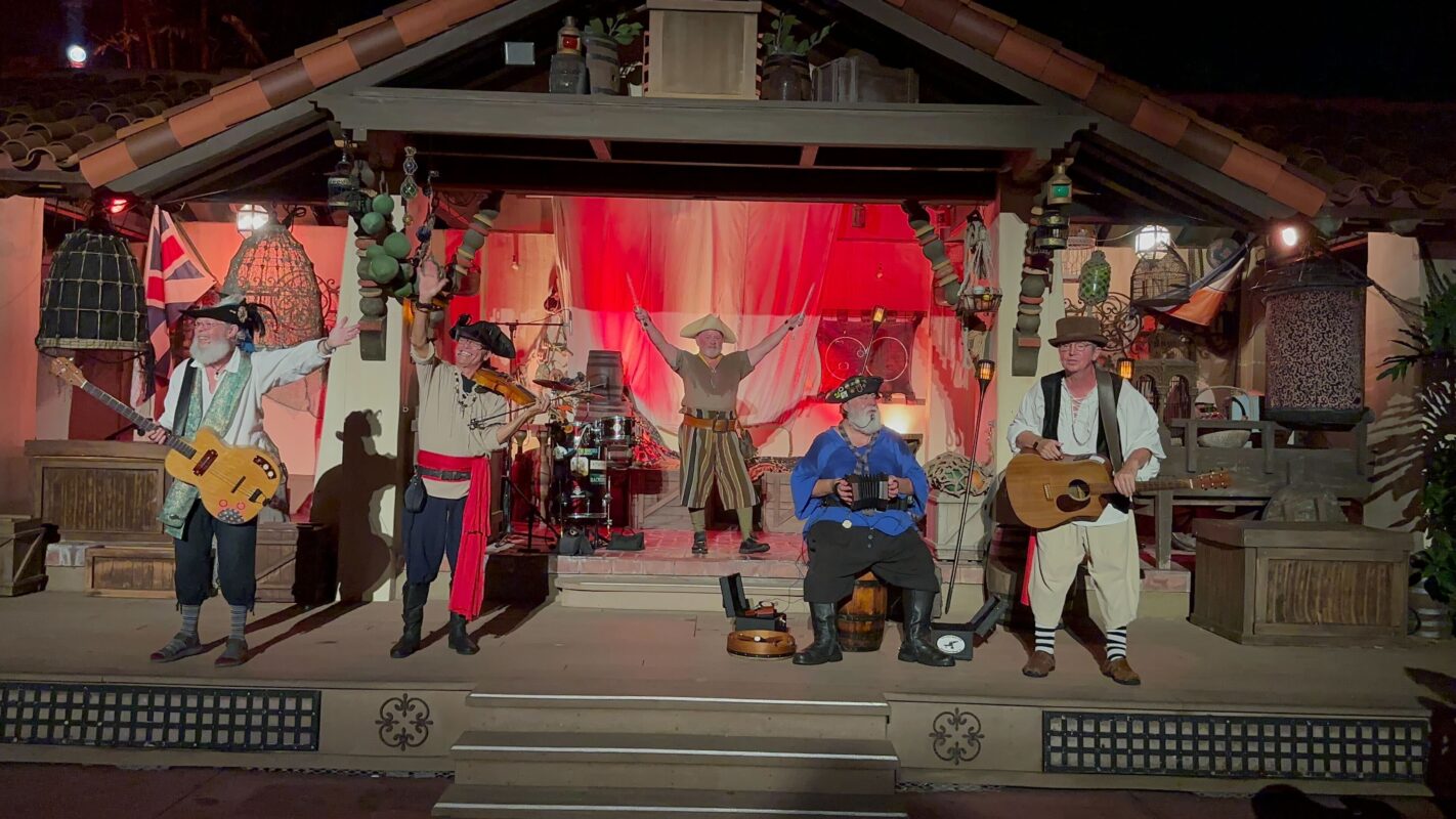 A band performs on stage dressed in pirate costumes, calling themselves "Rusty Cutlass." The setup includes various pirate-themed decorations and instruments, with a backdrop illuminated in red lighting.