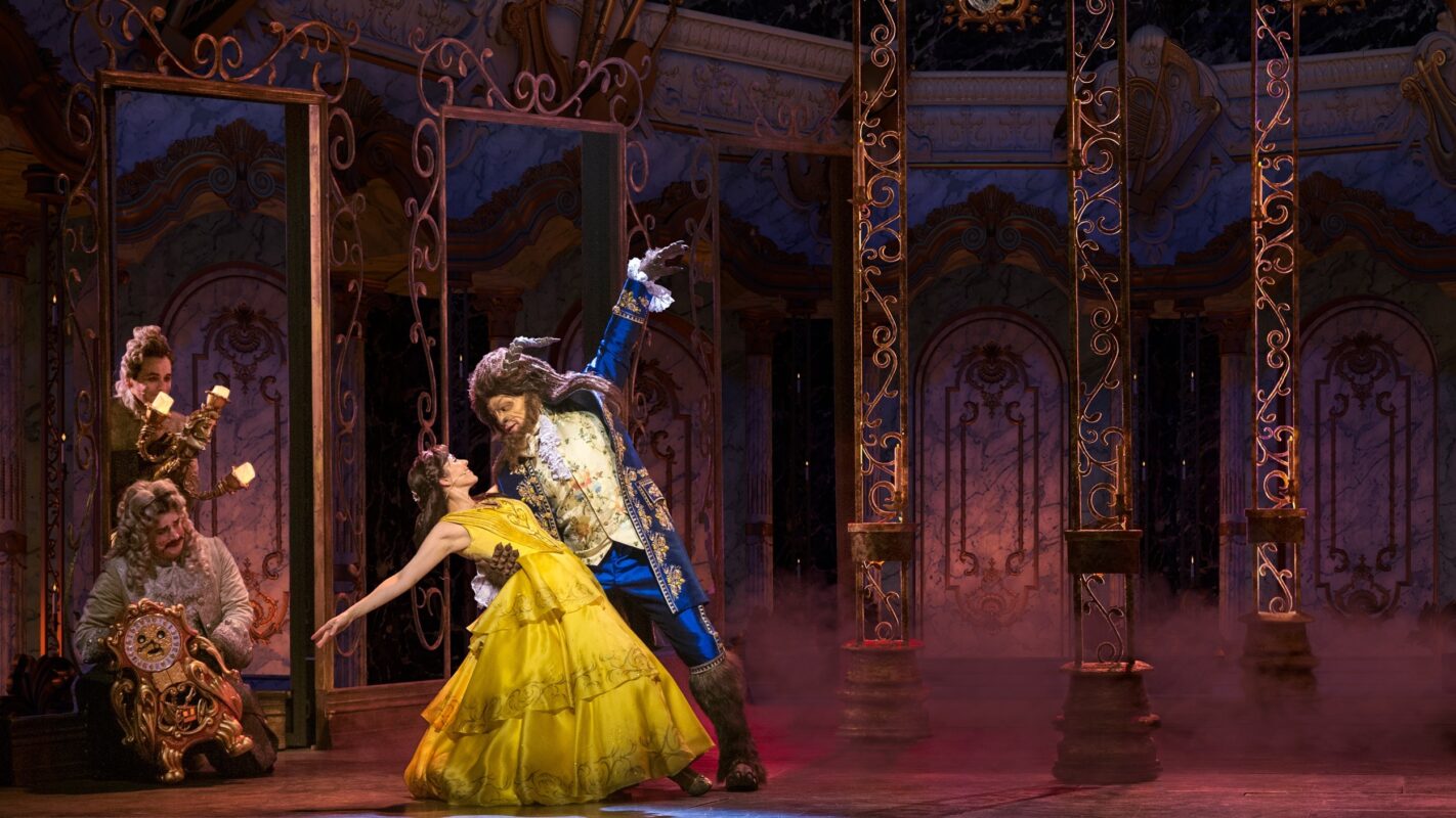 Actors perform a ballroom dance scene in an ornate, stage production. One actor, dressed as a beast, holds a woman in a yellow dress reminiscent of Disney treasure while others, adorned in period attire, look on from the background.