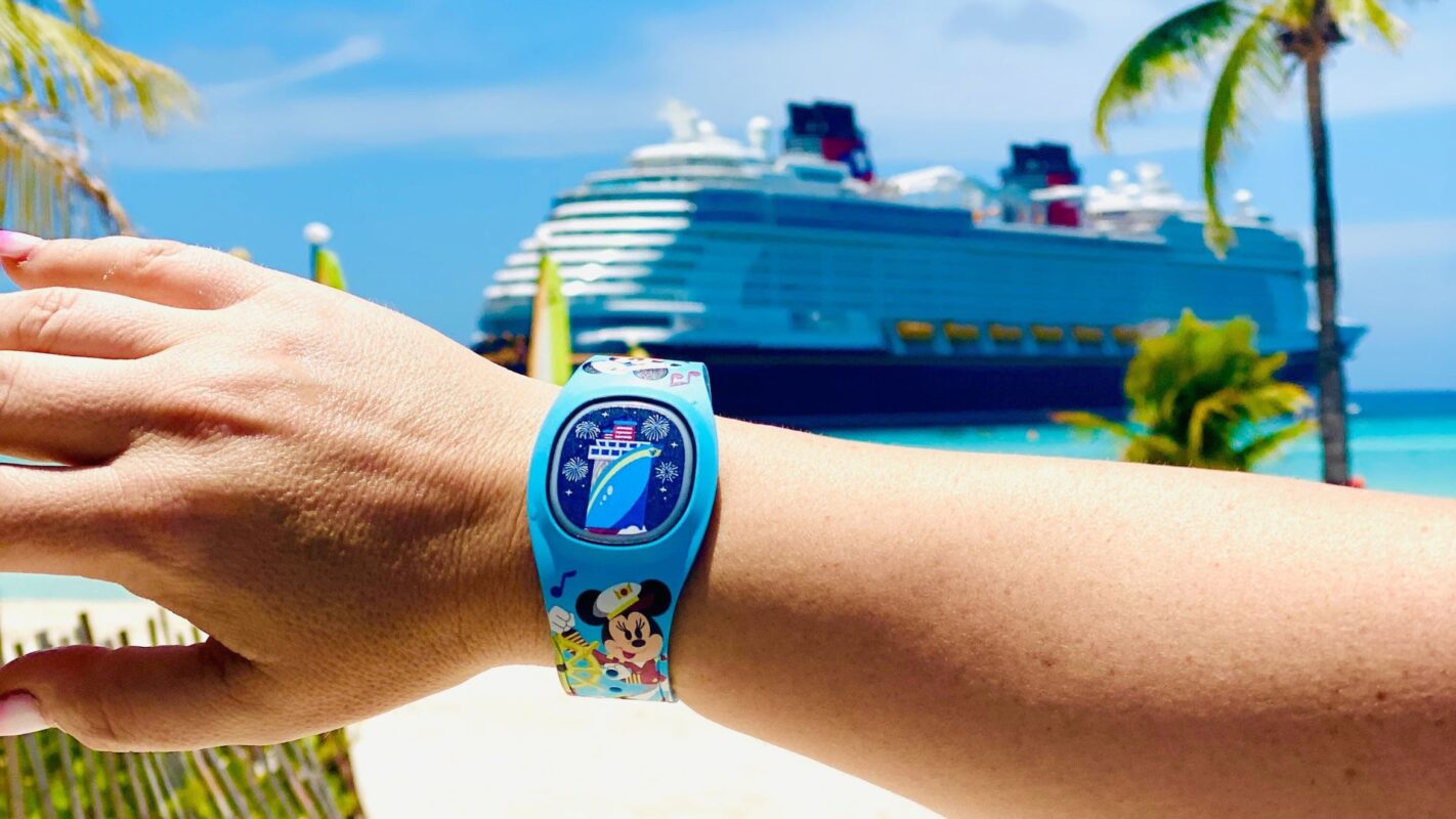 A hand wearing a Disney Cruise Line DisneyBand is held up against a backdrop of a cruise ship on a sunny beach with palm trees.