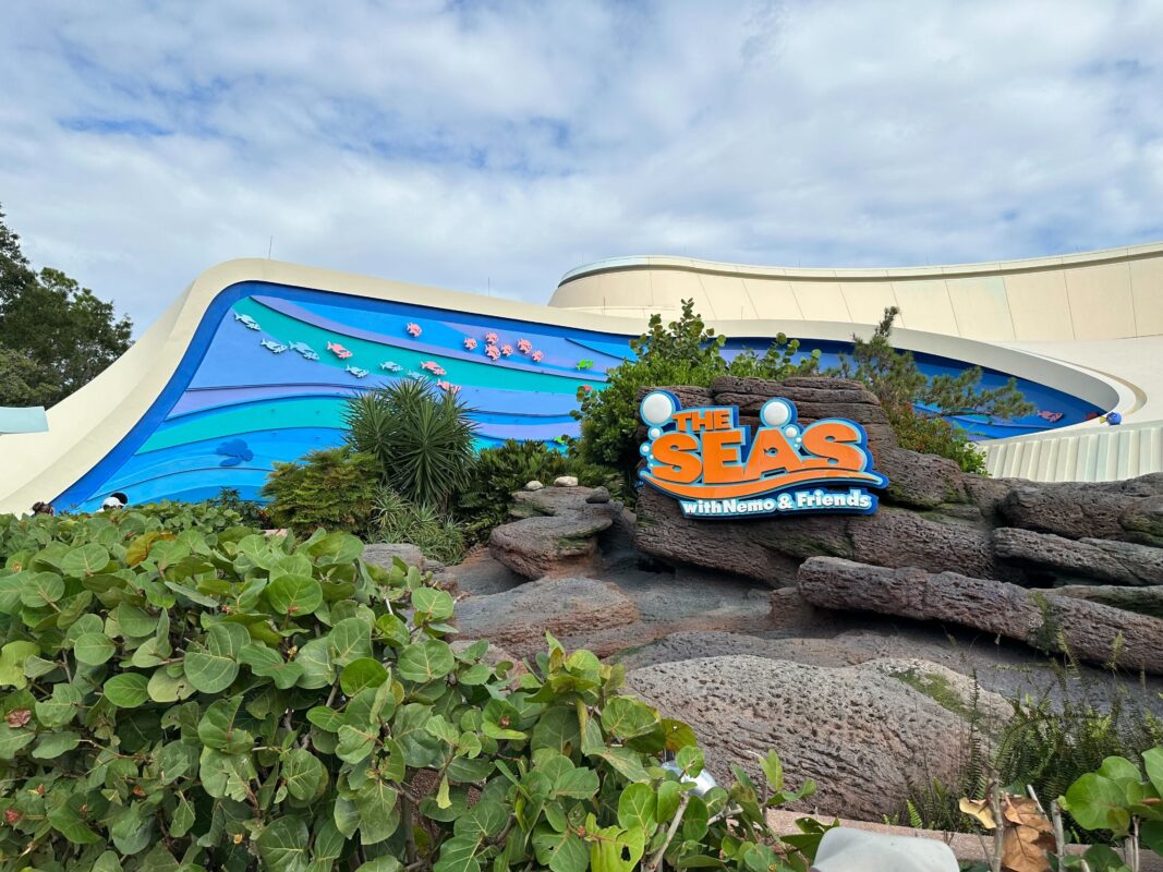 Exterior of The Seas with Nemo & Friends pavilion at Epcot, featuring an ocean-themed mural and a sign among rocks and greenery.