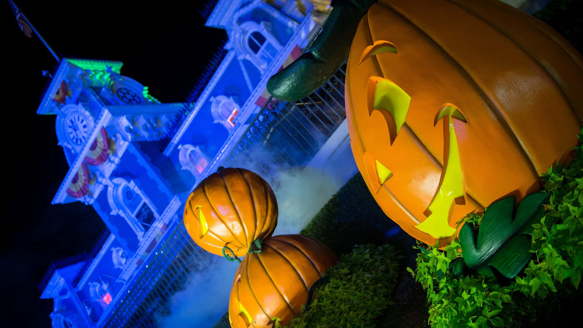 Large illuminated jack-o'-lanterns in front of a brightly lit, blue-hued building at night with light fog in the background, creating one of the scariest attractions around.