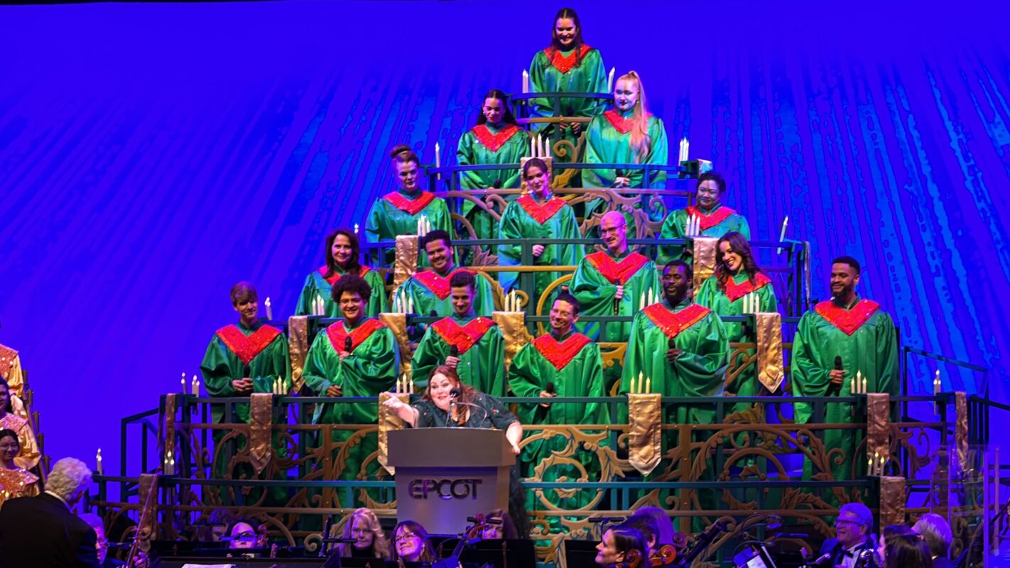 A choir in festive green robes with red accents performs on tiered risers at Epcot’s Candlelight Processional, while a conductor stands in front of the group.