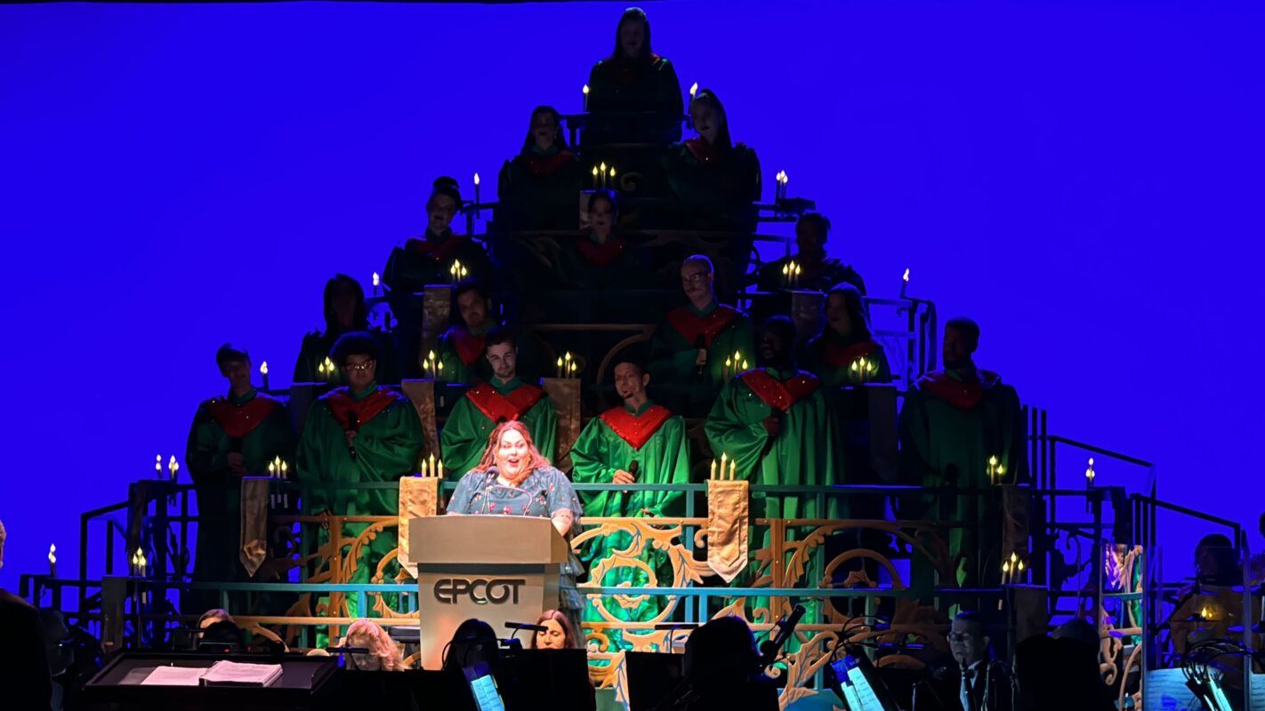 A speaker stands at a podium labeled "EPCOT," as part of the enchanting Candlelight Processional. Behind them, a choir in green robes and red scarves sings harmoniously against a blue backdrop, beautifully illuminated by glowing candles.