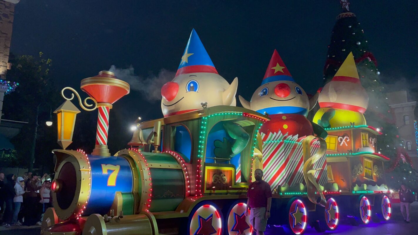 A festive parade float featuring a colorful, illuminated toy train with cartoon characters and holiday decorations moves through a night-time setting.