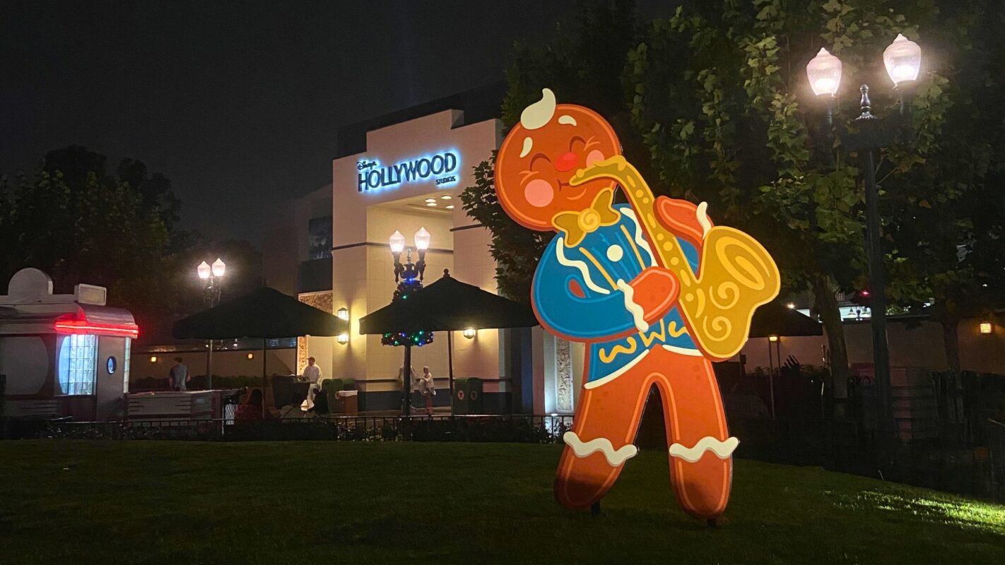 A large gingerbread man figure playing a saxophone stands on a grassy area in front of The Hollywood Lounge at night, illuminated by street lamps, perfectly capturing the festive spirit of Jollywood Nights.
