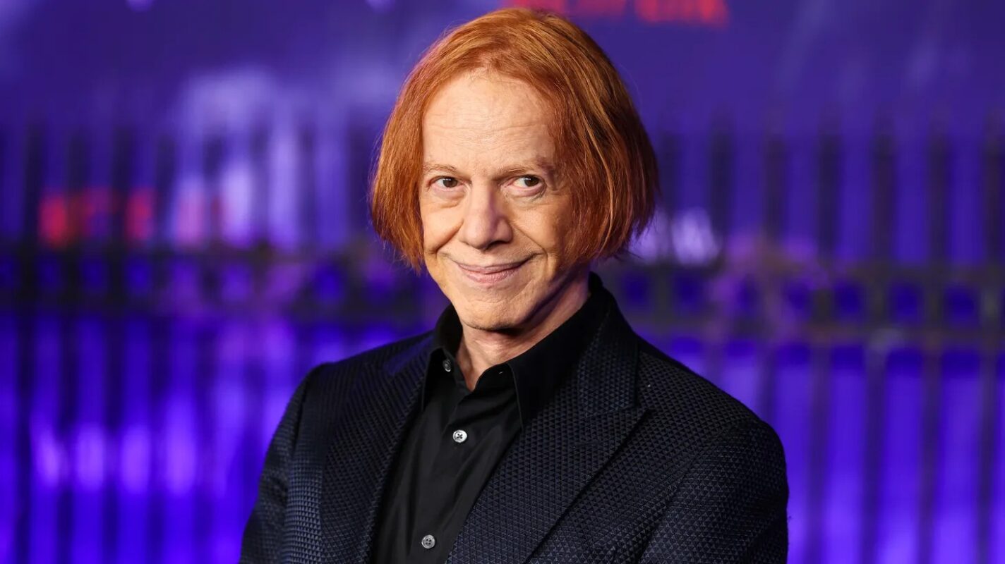 Danny Elfman, a man with red hair wearing a black suit, in front of a purple backdrop