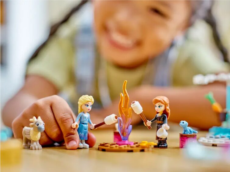 A child plays with their LEGO Disney mini dolls of Anna and Elsa, who are roasting marshmallows over a LEGO fire. Small figures of Bruni and a baby reindeer look on.