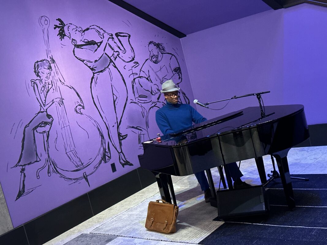 Joe Gardner, a Black man in a hat and blue top, playing piano in front of a purple mural of musicians