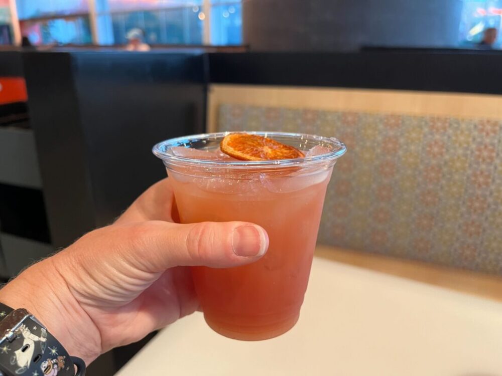 A clear plastic cup with a dark red cocktail and a blood orange slice on top.