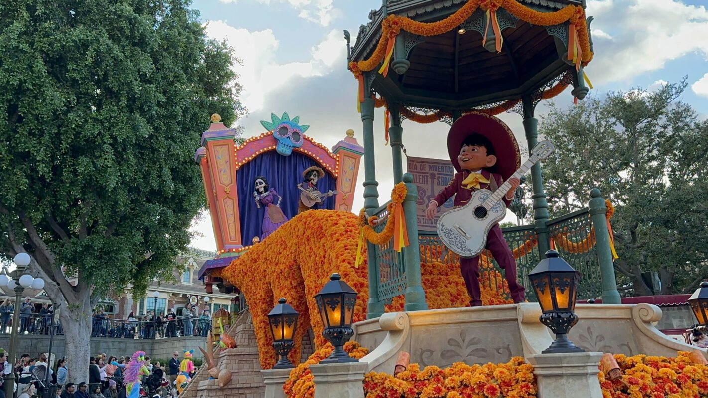 A colorful parade float features a character playing guitar, surrounded by vibrant orange marigolds. In the background, two skeleton figures are positioned on a stage where the magic happens. Spectators observe from the sidelines.