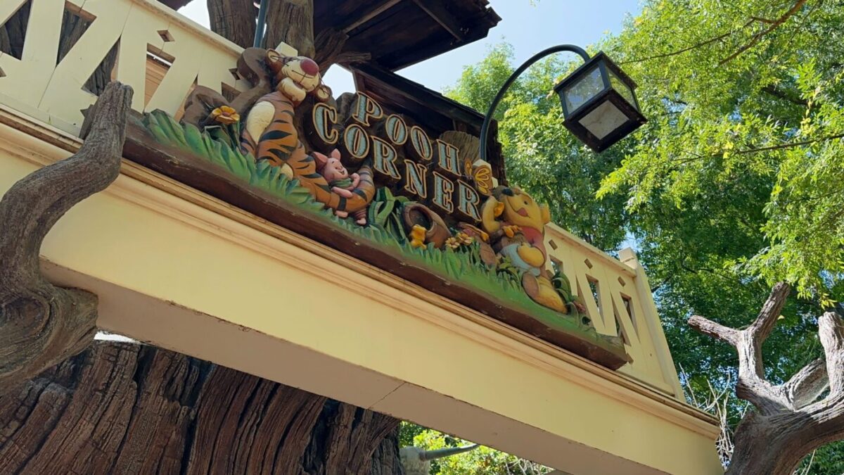 Signboard of "pooh corner" featuring colorful figures of winnie the pooh and friends, above a shop entrance, with a lamp post and trees in the background.