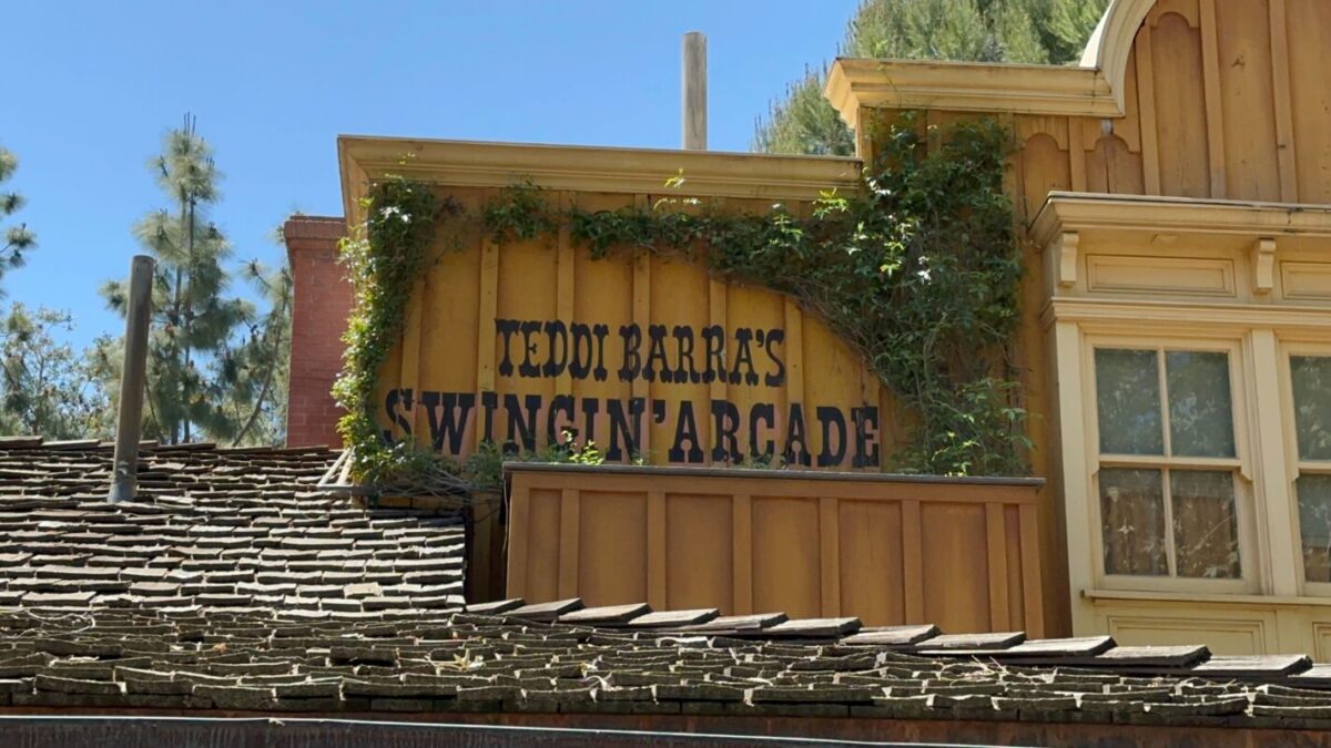 Sign reading "teddy barra's swingin' arcade" mounted on the wooden facade of a rustic building with a shingled roof.