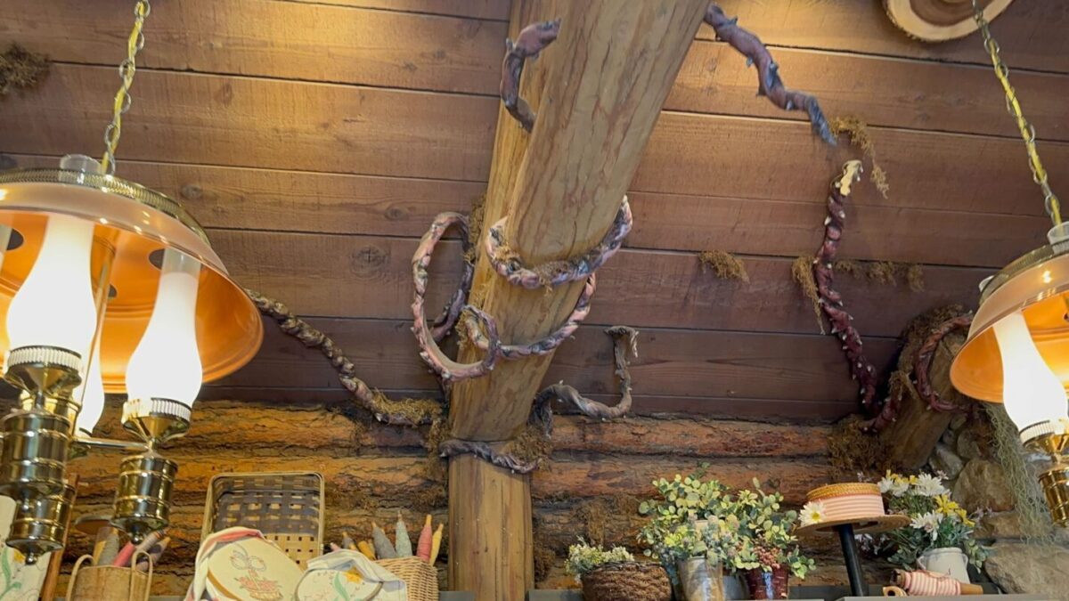 Interior of a rustic cabin with a wooden beam wrapped in twine, flanked by vintage orange lamps, against a backdrop of wooden walls adorned with floral baskets and trinkets.