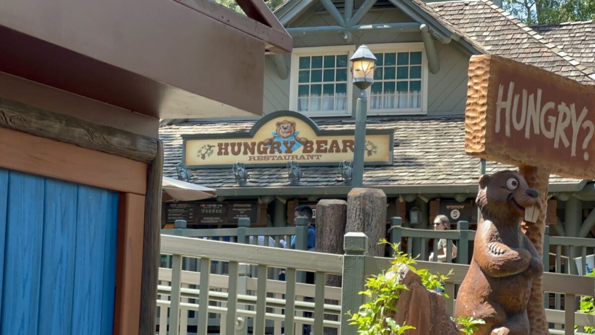 A wooden bear statue stands near the sign of the hungry bear restaurant in a theme park, with wooden fencing and a blue sky in the background.