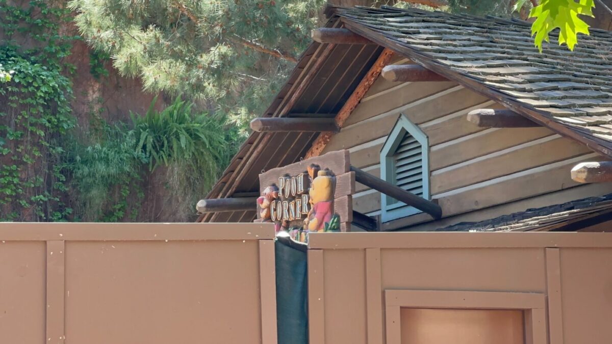 A cartoon-style sign with the text "pooh corner" on a rustic wooden cabin hidden partially behind a brown fence, surrounded by trees.
