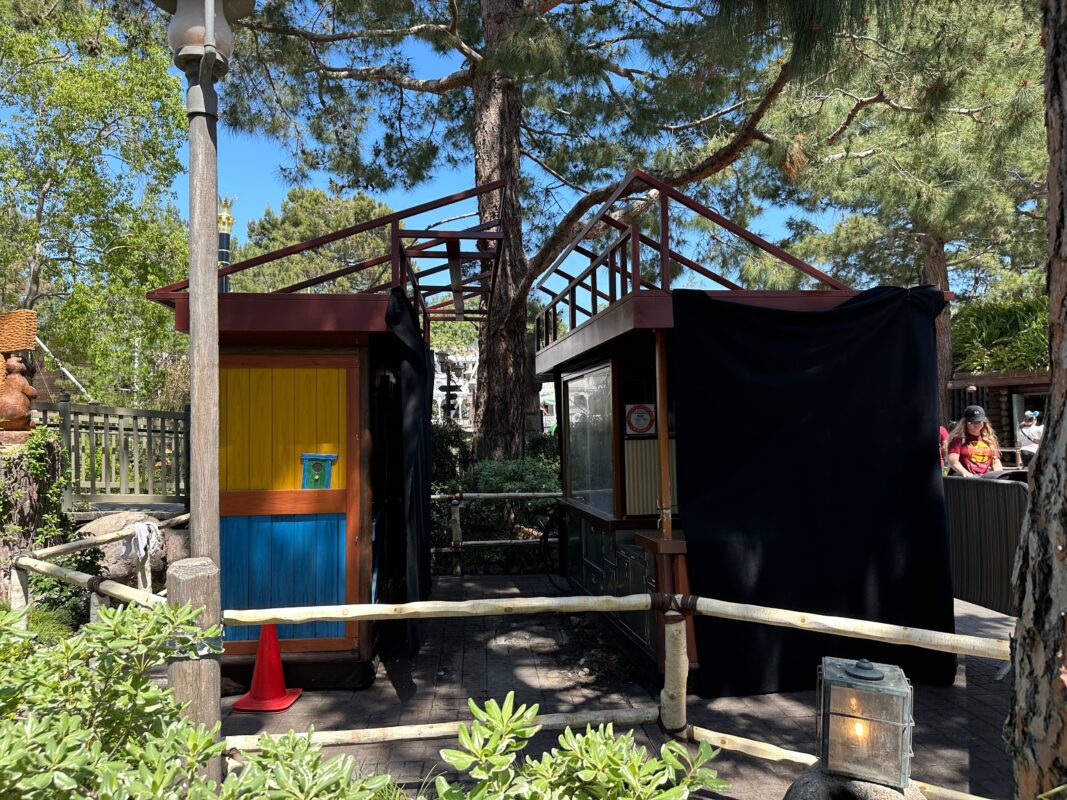 A colorful outdoor kiosk with a yellow door and a black curtain, surrounded by lush greenery and trees on a sunny day.