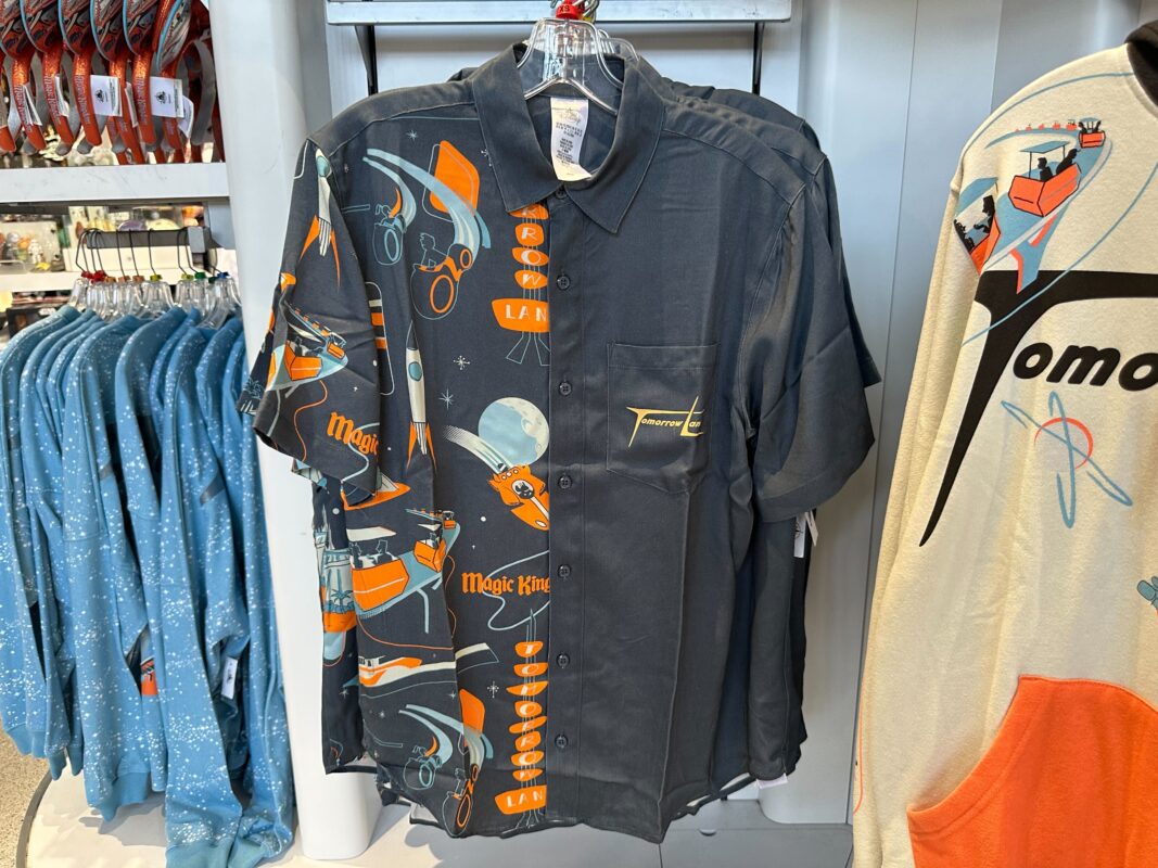 Black shirt with a bold, colorful print featuring retro-futuristic space designs, displaying "Magic King" text and various space-themed pictures, hanging on a rack next to other clothing items in a store. Nearby, you'll find the Tomorrowland ear headband that perfectly complements this celestial style.
