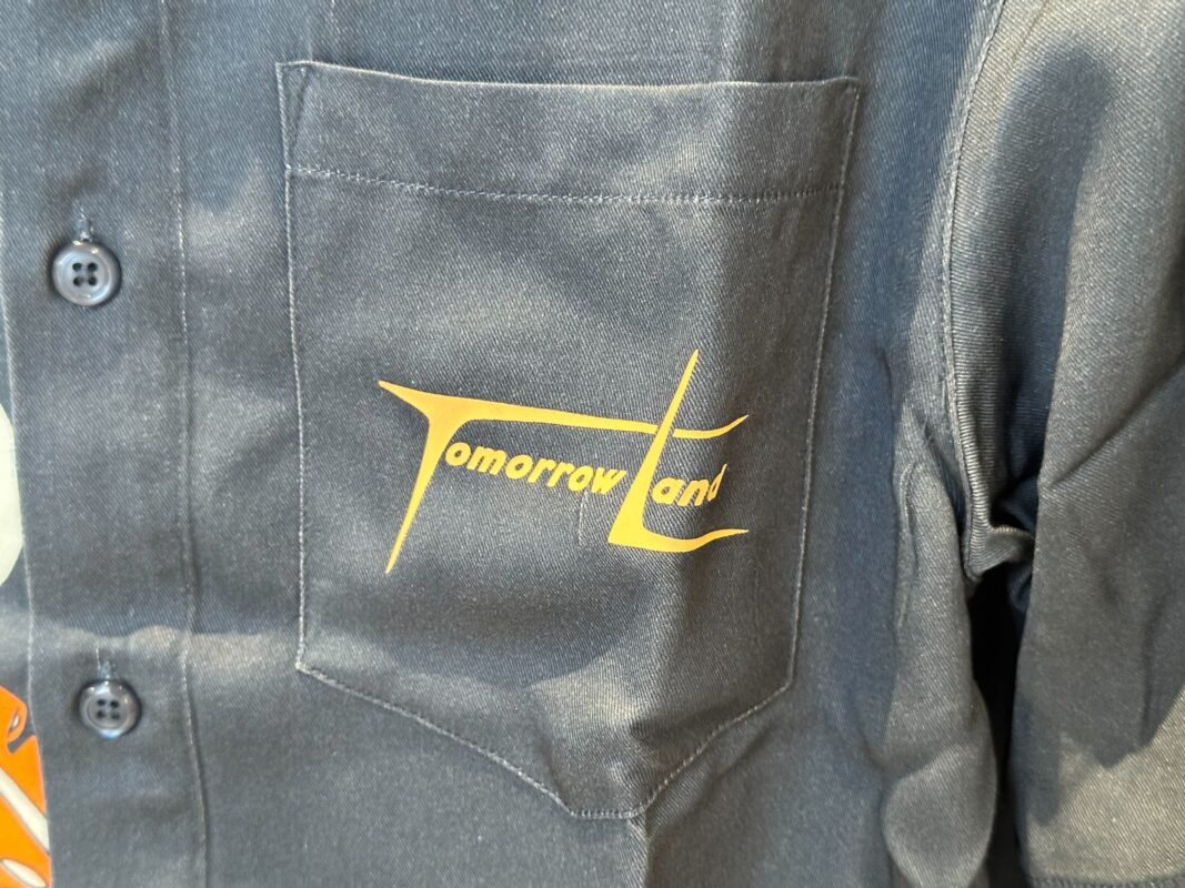 Close-up of a dark-colored shirt with a pocket on the chest. The pocket has the text "TomorrowLand" in yellow print, matching perfectly with the Tomorrowland ear headband.