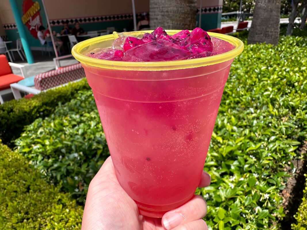A hand holding a plastic cup filled with a vibrant pink slush drink, topped with chunks of pink ice, against a green shrubbery background.