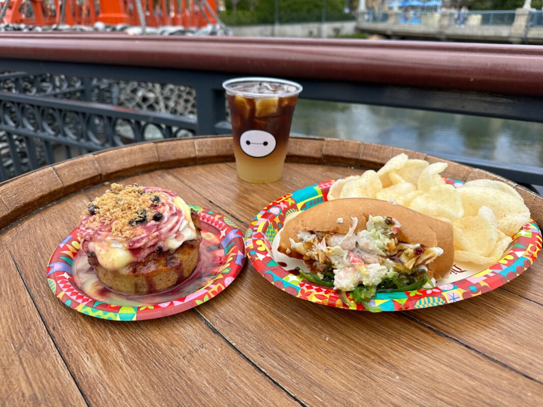 A meal at Aunt Cass Café on a wooden table featuring a berry-topped muffin, a sandwich with chips, and a cold brew coffee, served on colorful plates.