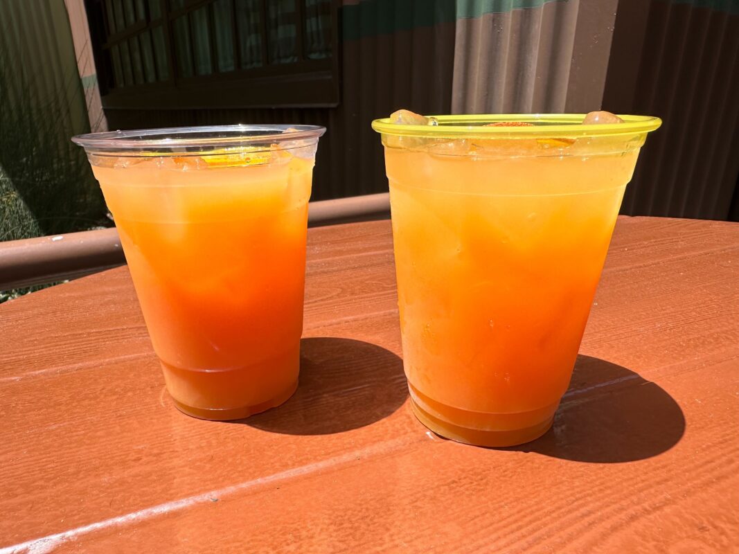 Two cups of orange juice with ice, one covered with a green lid, standing on a wooden table in sunlight.