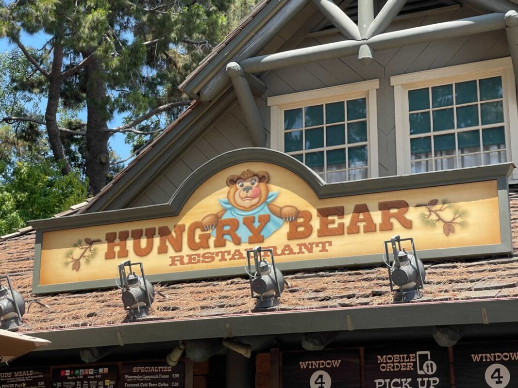 The image shows the exterior of Hungry Bear Restaurant in New Orleans Square, a building with a sign displaying a cartoon bear holding a spoon and fork. There are order windows and a menu below the sign.