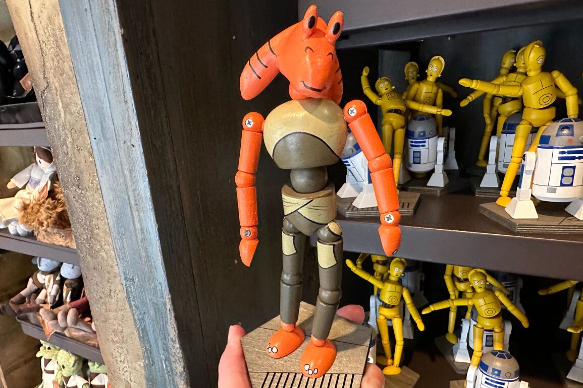 A hand holds an orange and brown robot toy resembling a character, displayed in front of a shelf with other figurines, including R2-D2 and C-3PO.
