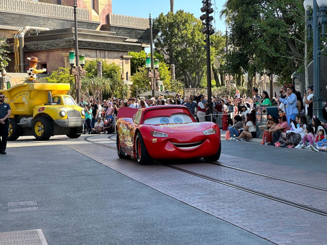 A red animated character car, resembling Lightning McQueen, parades down a street lined with spectators during the Pixar Pals Celebration at Disney California Adventure, followed by a large yellow truck. Trees and a building are visible in the background.