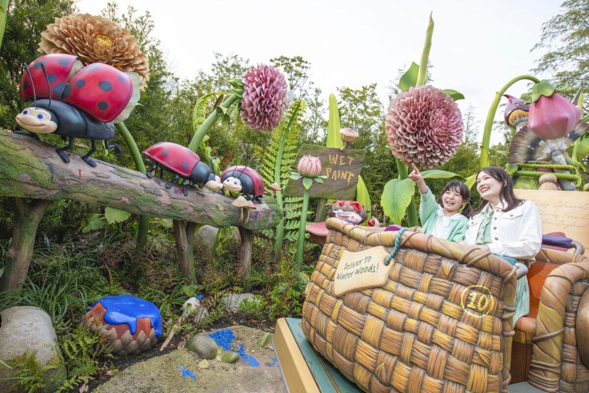 Two people in a themed amusement park ride smiling and pointing at oversized, colorful models of insects and flowers.