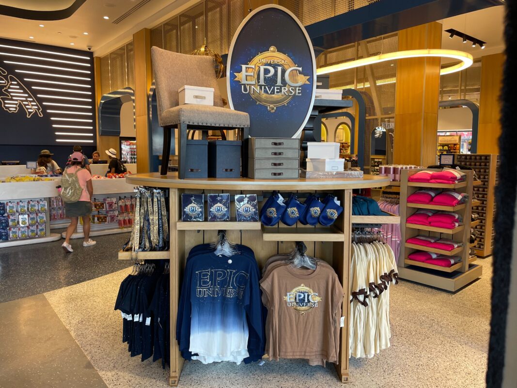 Epic Universe merchandise display at Universal Studios Store with assorted apparel including t-shirts and hats, and themed decorations.