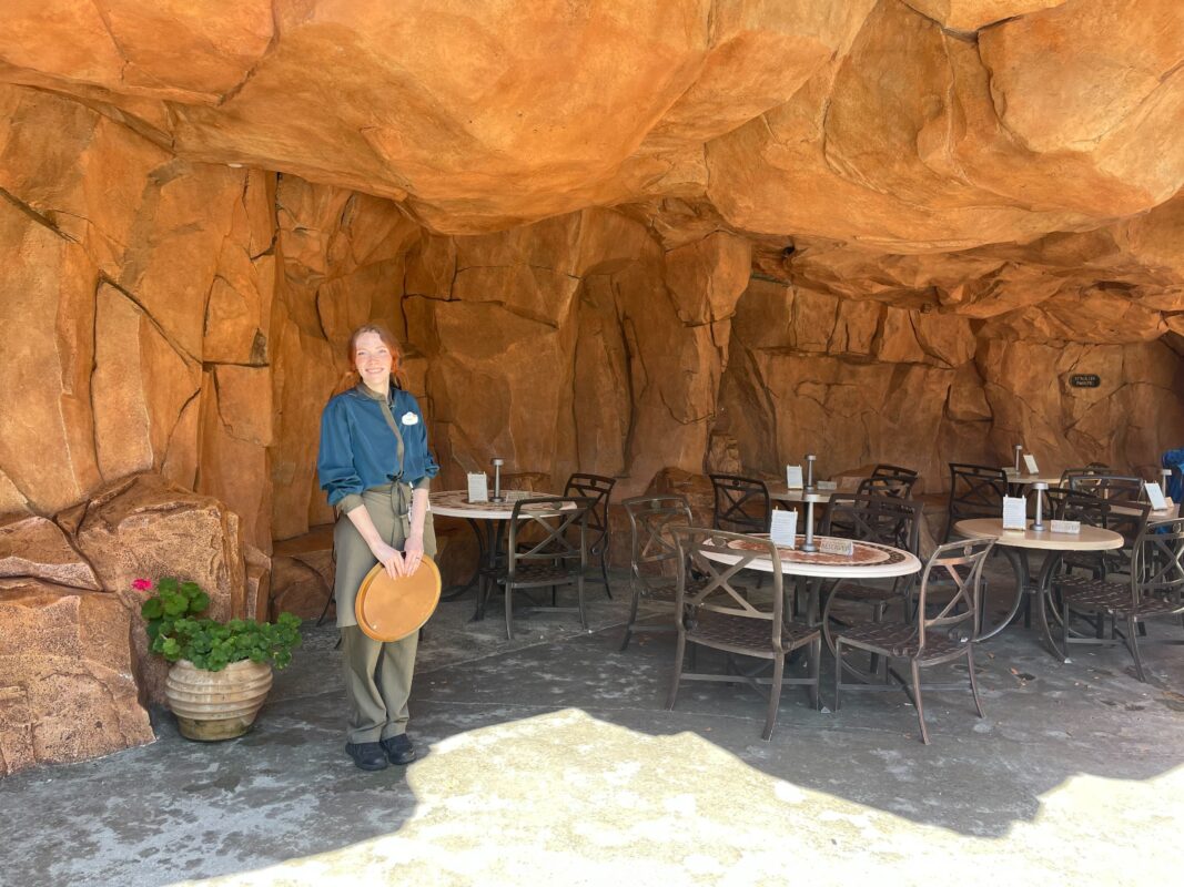 A person in a uniform stands in front of outdoor tables set against a rock-like wall at the Mythos Restaurant. Nearby, a plant adds greenery to the lounge area, which features a few empty tables and chairs, perfect for enjoying Dueling Dragons Mocktails.