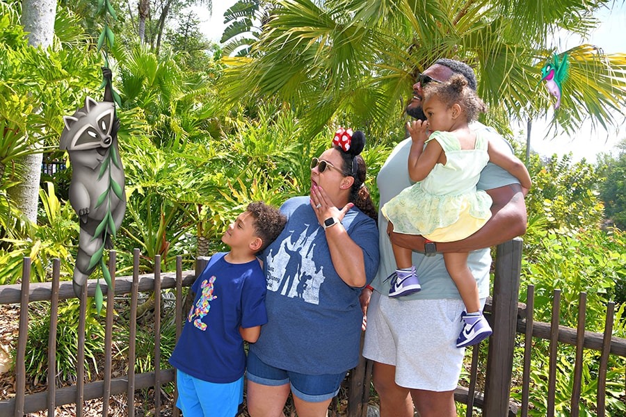 A family of four stands outdoors near a fence, looking towards an animated character in a tropical setting. The man holds a young girl, while the woman stands beside a boy wearing a blue t-shirt.