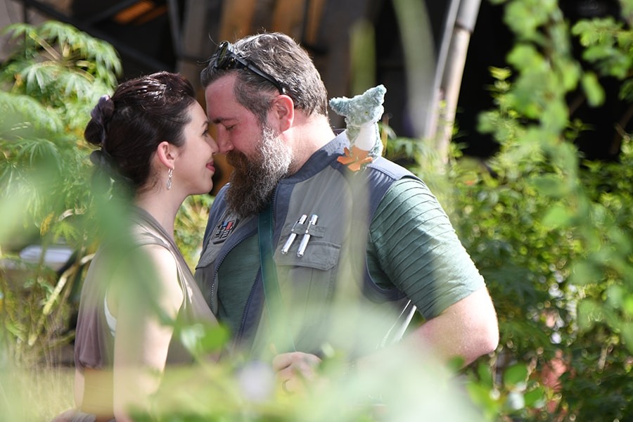 A man and woman stand close together outdoors among greenery. The man wears a vest with pens in the pocket and sunglasses on his head, while the woman smiles with her eyes closed, touching foreheads. This sweet moment captures their excitement for new opportunities ahead, perfect for a PhotoPass memory.