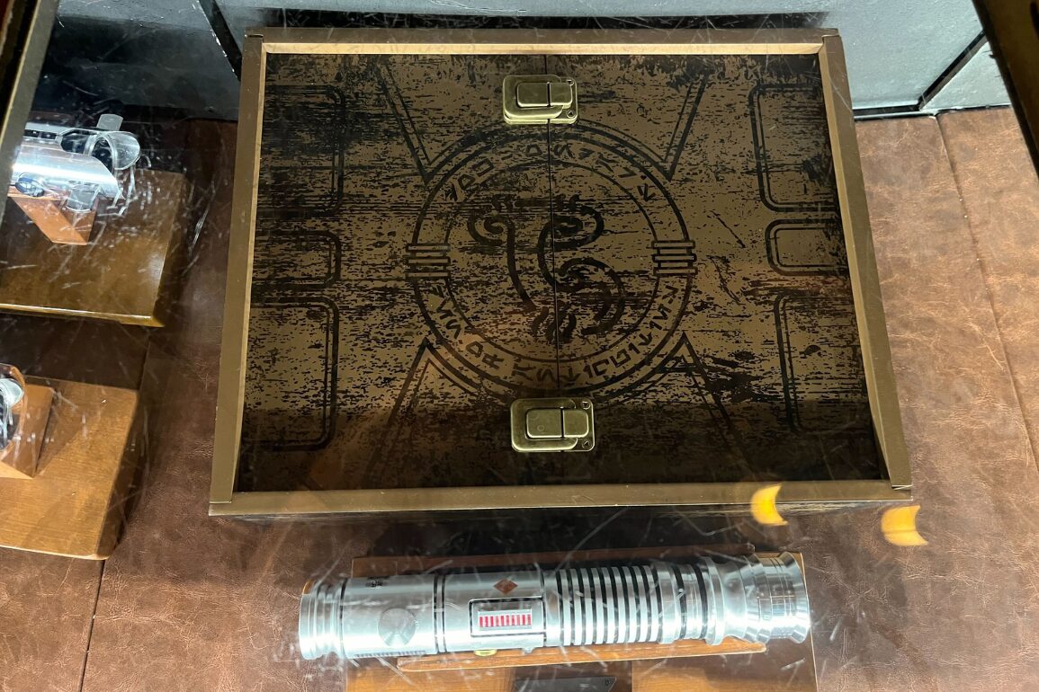 Wooden box with intricate designs and metallic clasps, displayed above a silver cylindrical object, possibly a part of a tool or device, on a brown surface.