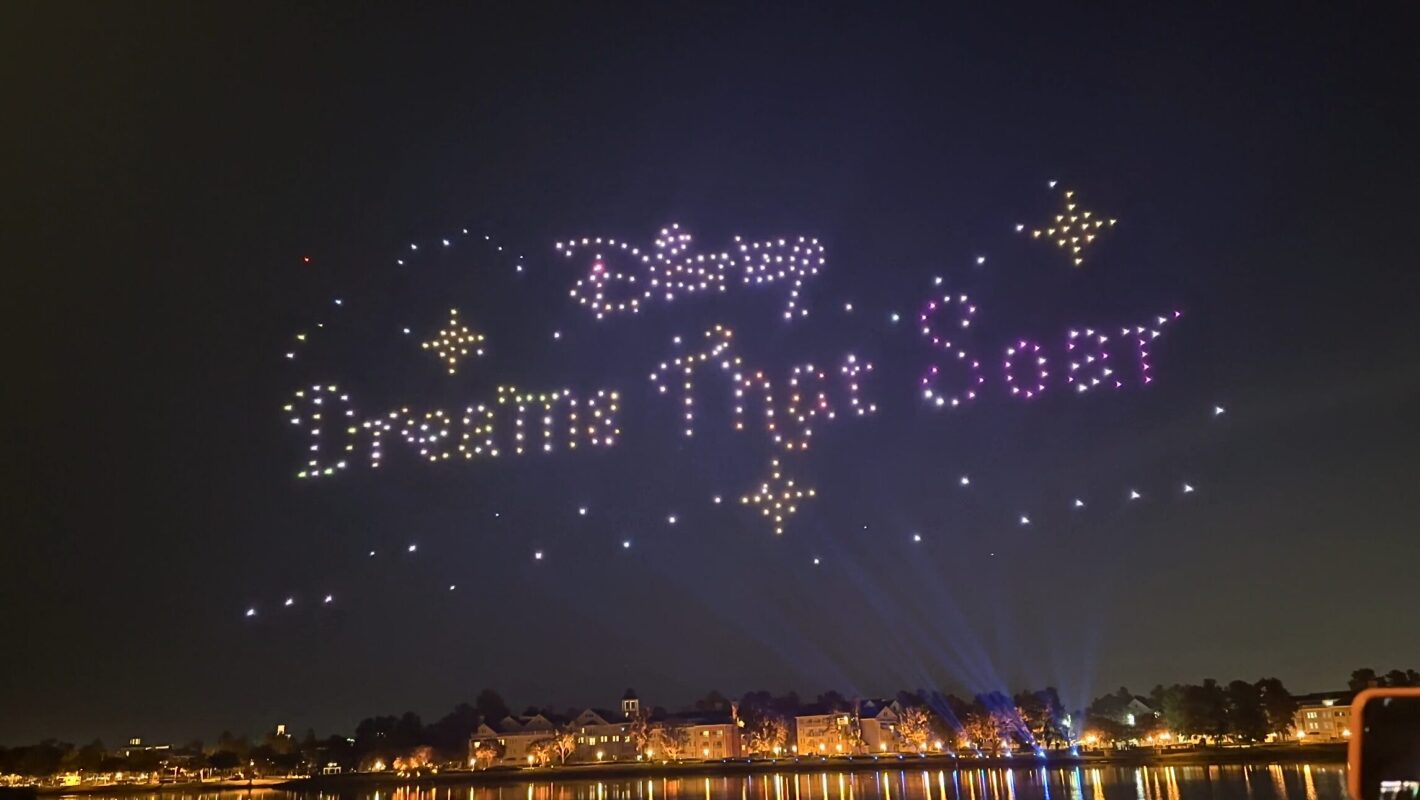 A nighttime sky display shows the words "Disney Dreams That Soar" formed by illuminated drones, with a lake and lit buildings visible below.