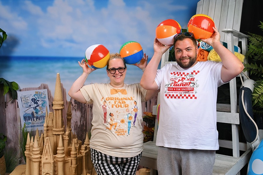 Two people stand in front of a beach-themed backdrop at Walt Disney World, each holding two colorful beach balls over their heads. They are smiling, and a sandcastle model is visible in the foreground, creating new PhotoPass opportunities for guests to capture the perfect vacation moment.