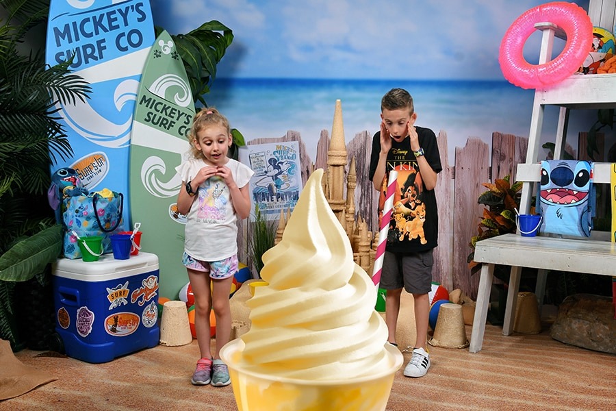 Two children stand in front of a large, swirled soft-serve ice cream cone in a tropical beach-themed setup at Walt Disney World, with surfboards, beach toys, and colorful decorations around them. It's one of those perfect PhotoPass photo opportunities you'll cherish forever.