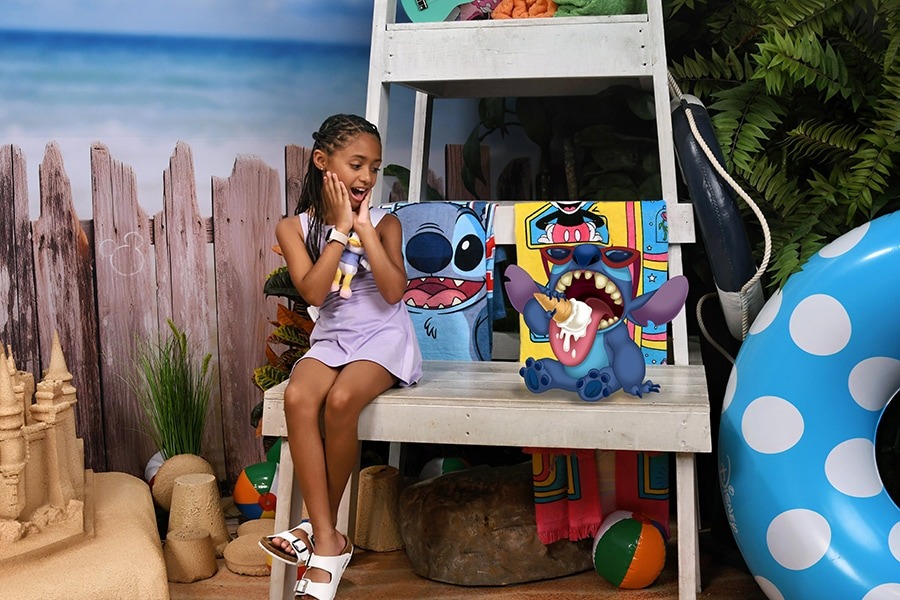 A young girl in a purple dress sits on a wooden bench, smiling at cartoon character Stitch, who is holding an ice cream cone and sitting next to her. The beach-themed background with sand and decor makes for one of the perfect PhotoPass opportunities at Walt Disney World this summer.