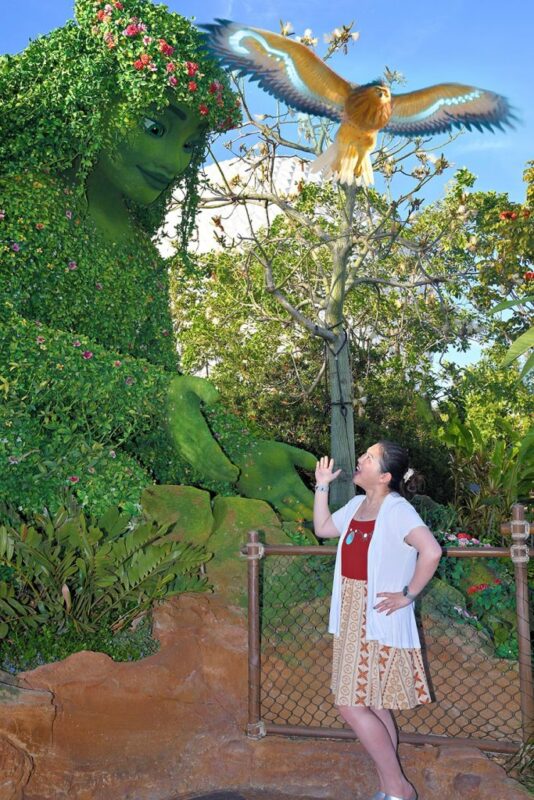 A woman stands posing with her hand extended near a large green plant figure as a bird flies above in the lush vegetation, all set under a clear sky. This summer, discover unique PhotoPass opportunities to capture your unforgettable moments, just like at Walt Disney World.