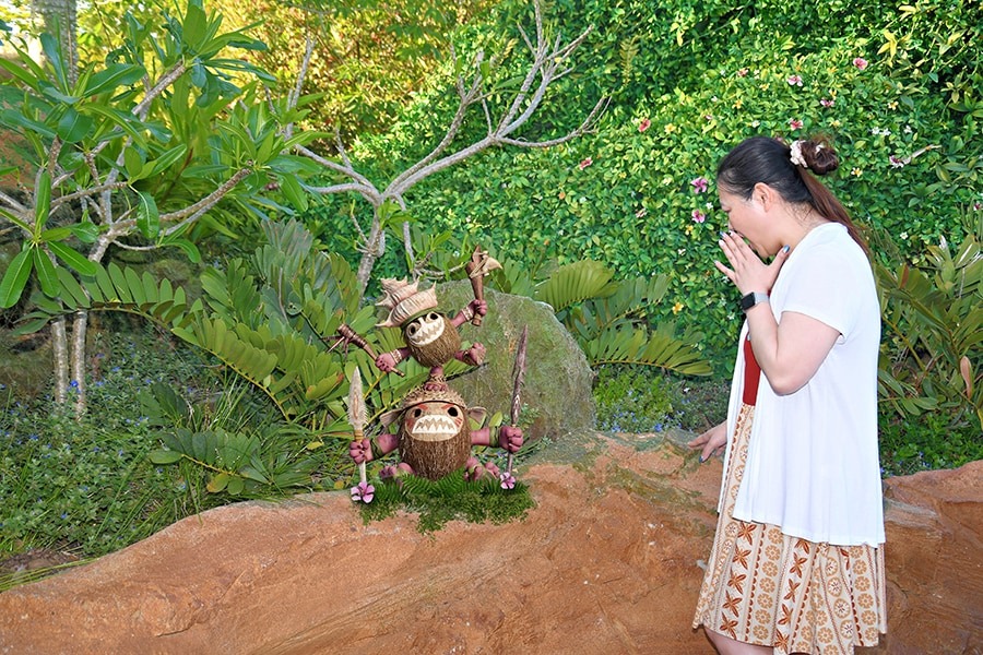 A woman stands next to a small statue of two stacked coconuts with faces and spears, set in a garden with lush greenery at Walt Disney World.