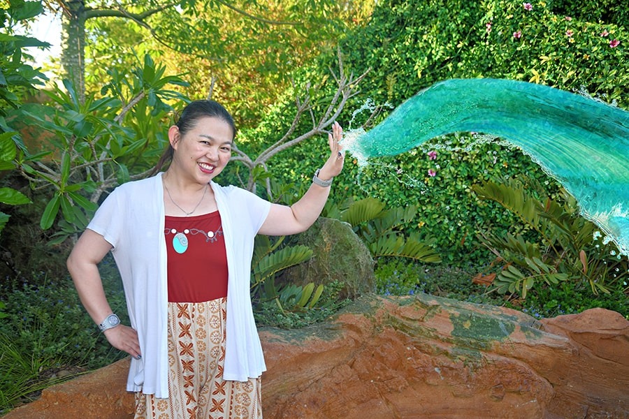 A woman in a white cardigan and red top stands next to a water-like green sculpture in a lush, outdoor setting. She is smiling and touching the sculpture with her right hand, perfectly capturing the magic of summer at Walt Disney World.