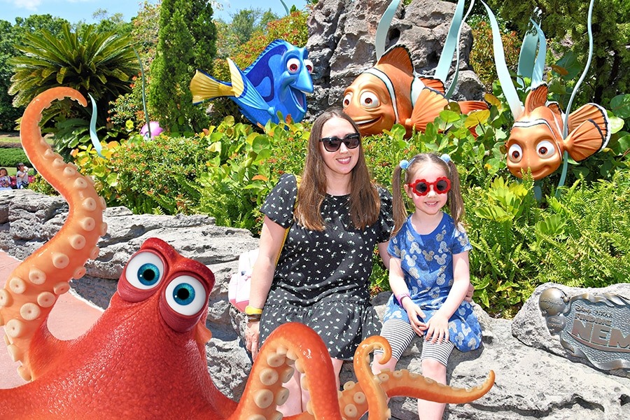 A woman and a young girl sit on a rock surrounded by statues of characters from Finding Nemo, including Dory, Marlin, Nemo, Pearl, and Hank. With vibrant greenery in the background, this enchanting PhotoPass moment captures the magic of Walt Disney World perfectly in the summer.