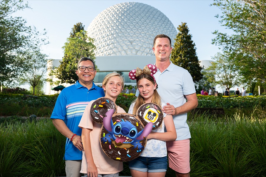 Four people pose in front of a large white geodesic dome at Walt Disney World. Two of them hold large Stitch-themed doughnuts, capturing a perfect summer memory. It's one of those quintessential PhotoPass opportunities you won't want to miss!