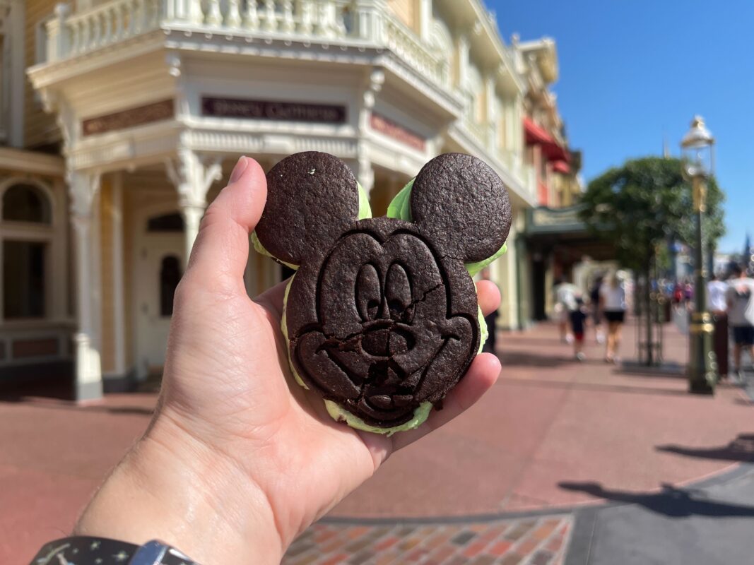 A hand holds a Mickey Mouse-shaped ice cream sandwich in front of a street with buildings and people walking in the background.