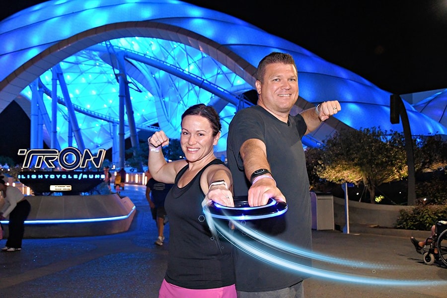 Two people pose with light cycle handlebars in front of the blue-lit 'TRON Lightcycle / Run' attraction at night, capturing a perfect memory at Walt Disney World with one of the best PhotoPass opportunities.