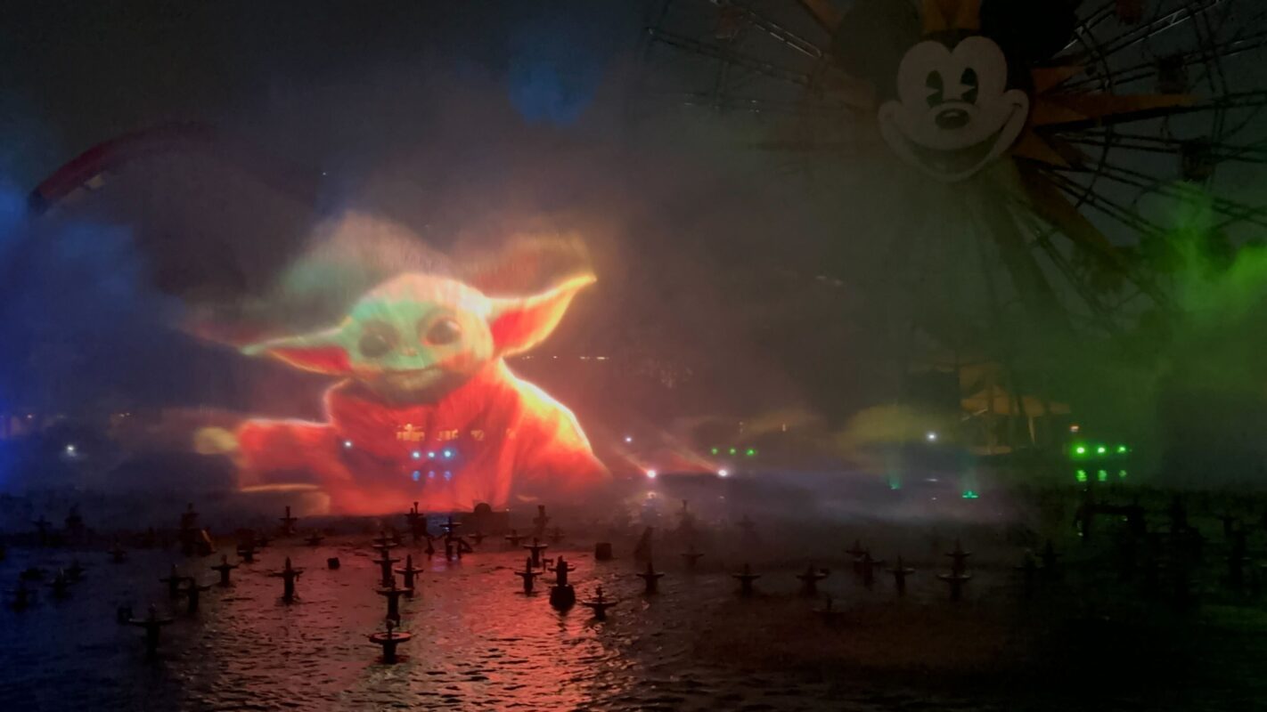 A large water projection of a character resembling Baby Yoda is displayed on a misty water surface at night, with an amusement park Ferris wheel featuring Mickey Mouse in the background.