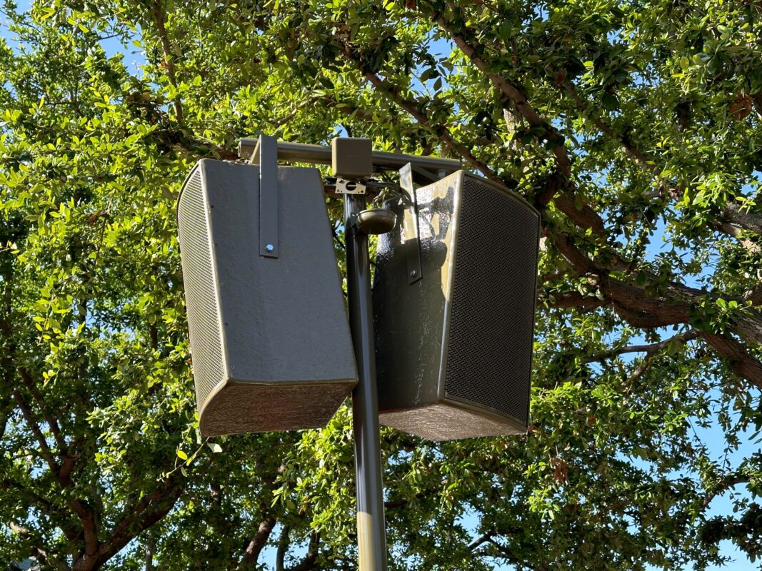 A pair of outdoor speakers mounted on a pole with green tree foliage in the background.