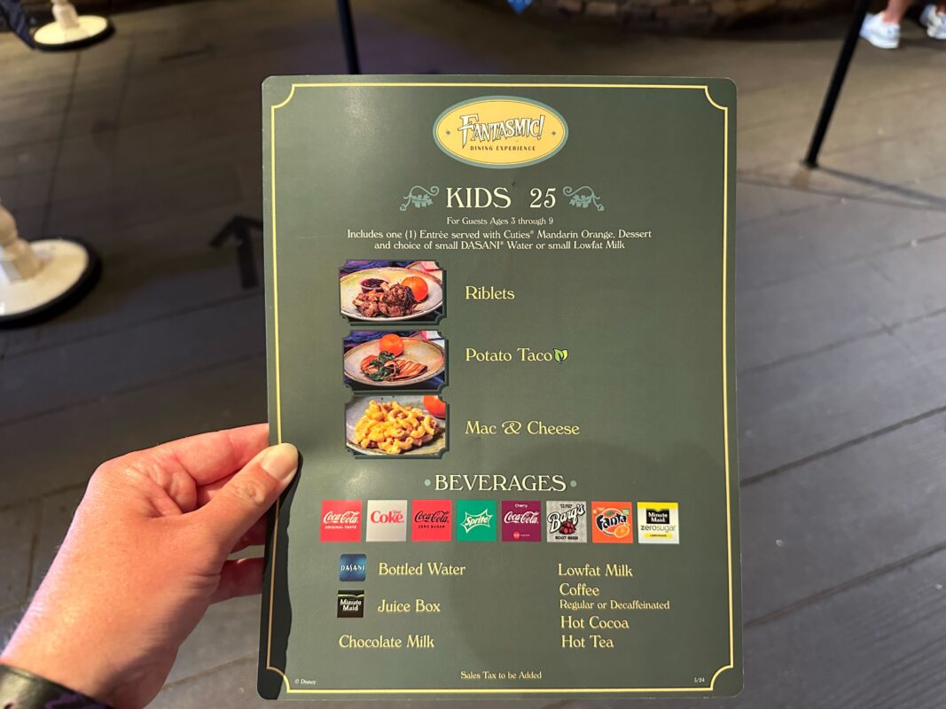A hand holds a kids' menu from a restaurant. The menu lists riblets, potato tacos, and mac & cheese as main options, with beverages including various sodas, bottled water, juice box, chocolate milk, and more.